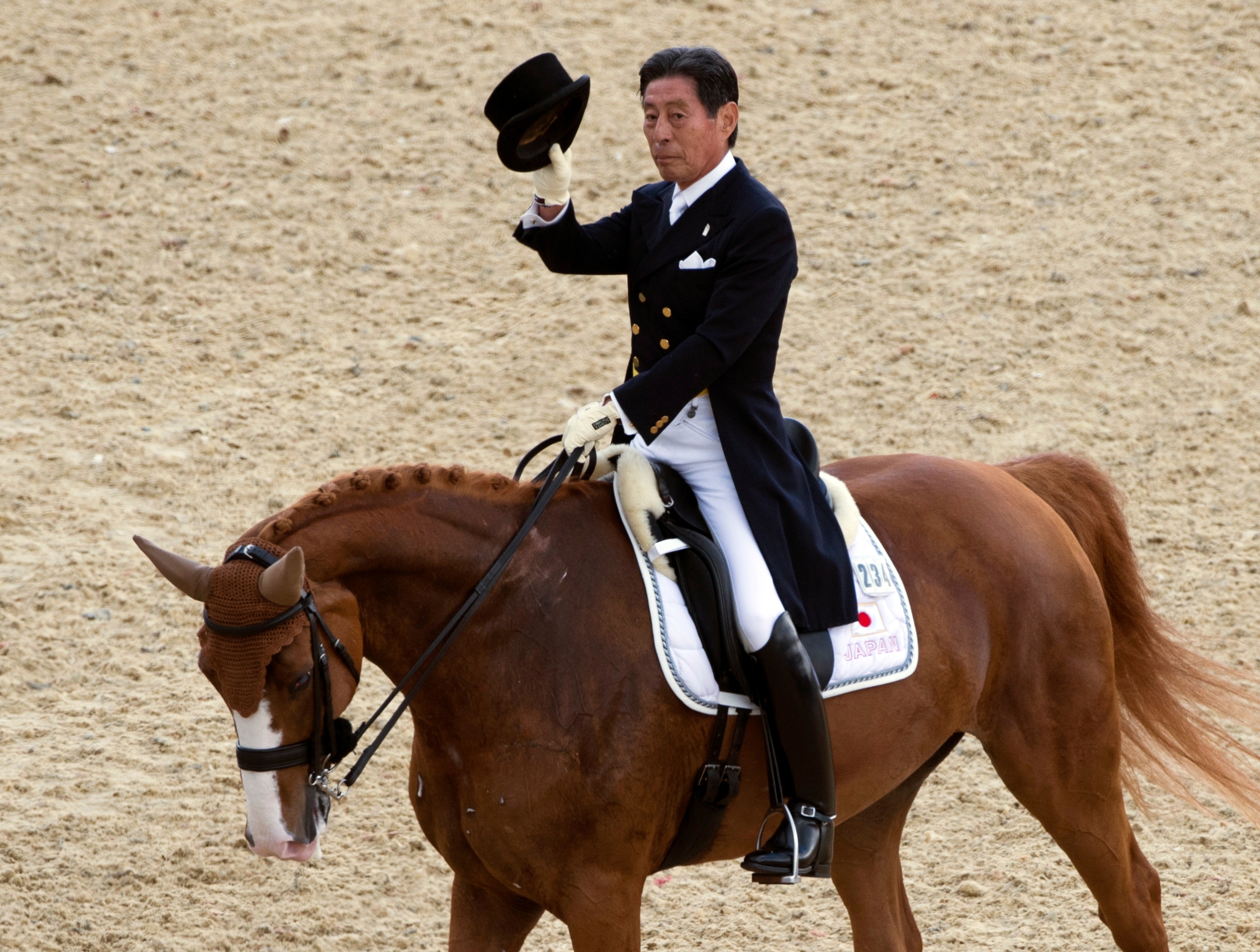 Person in formal equestrian attire on a horse, holding a top hat in a gesture, on a sandy arena