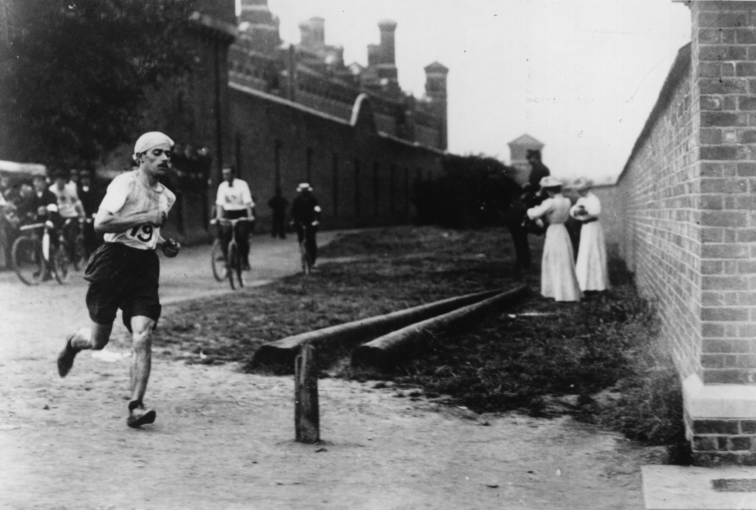 Runner in early 20th-century marathon near brick walls, surrounded by cyclists and onlookers