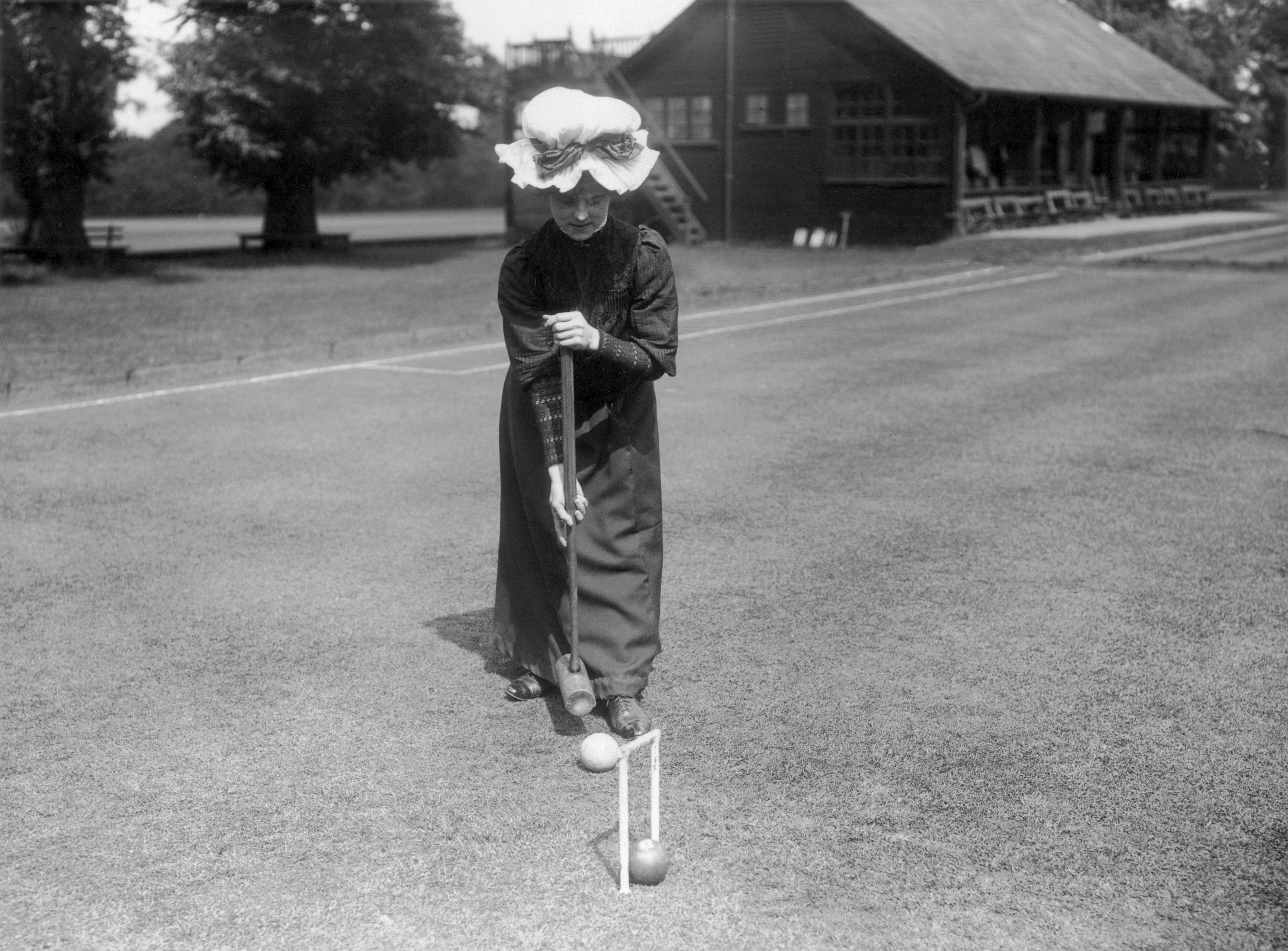 A vintage photo of a woman in an old-fashioned dress and hat playing croquet on a grassy field near a wooden clubhouse