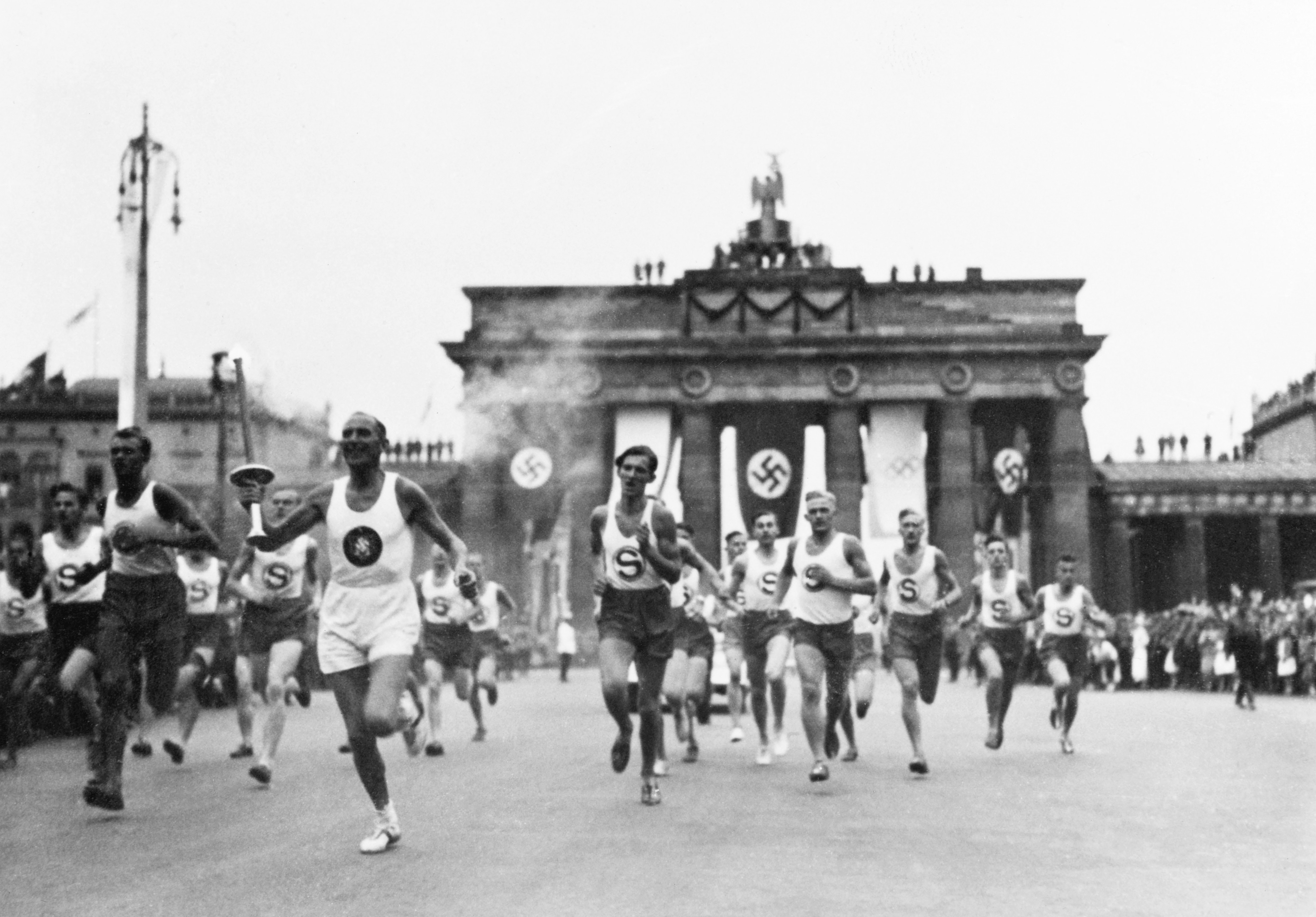 Athletes running in a torch relay near the Brandenburg Gate in Berlin, 1936, featuring Nazi symbols prominently displayed