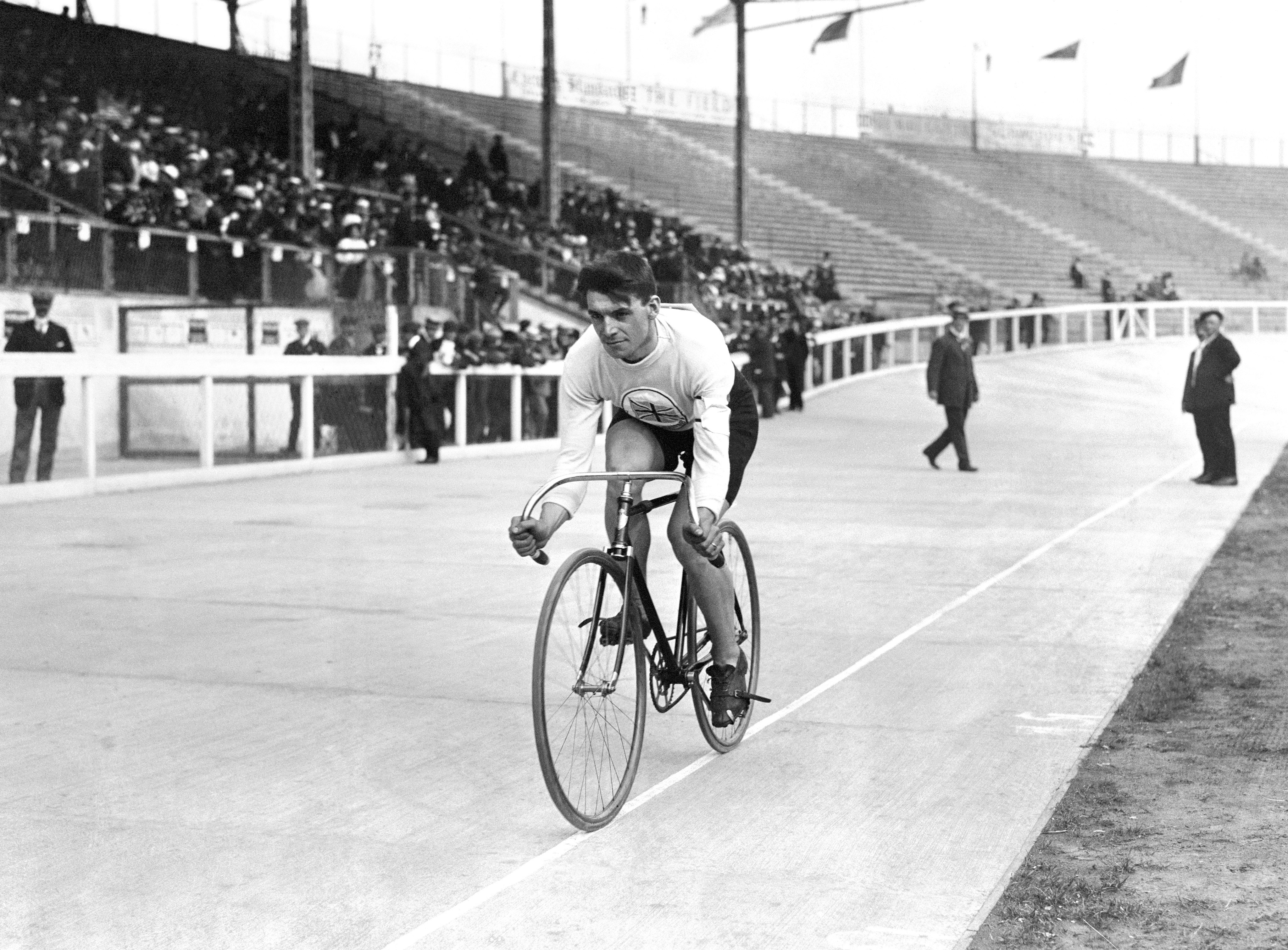 Cyclist in an early 20th-century velodrome race, leaning forward on a single-speed bike, with spectators in the stands