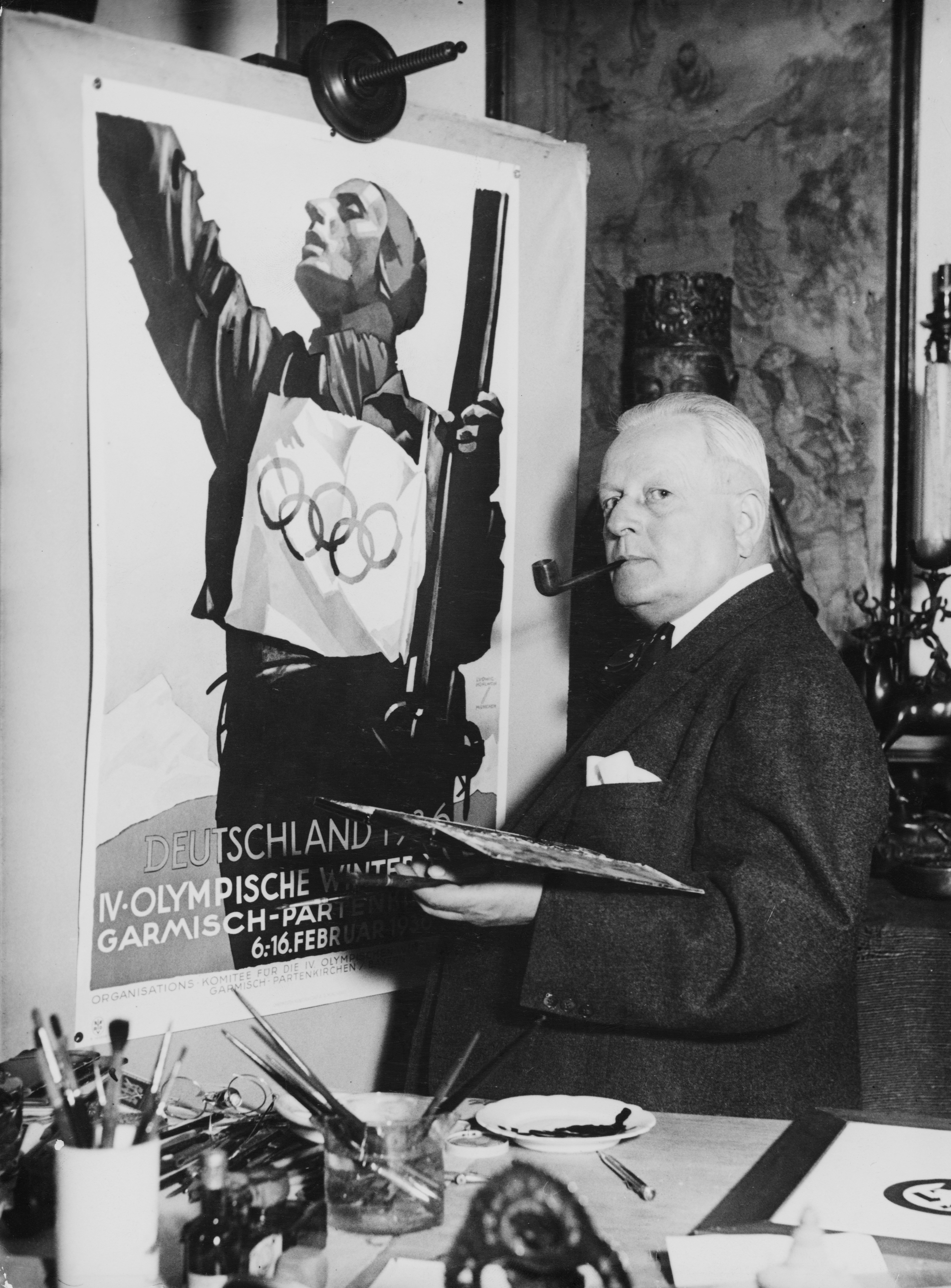 An artist with a pipe stands beside an Olympic poster he is painting, featuring a skier. The setting suggests a studio environment