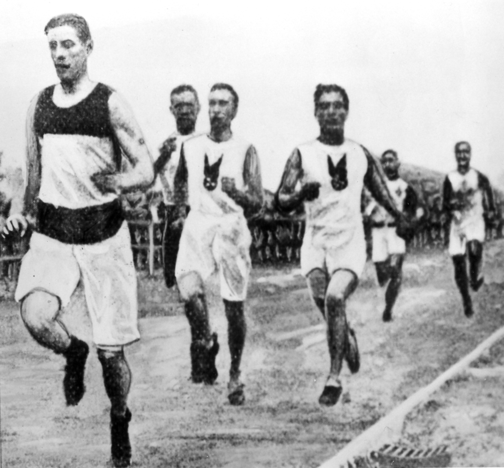 Historic black-and-white photo of male athletes running in a race on a road, wearing vintage athletic uniforms