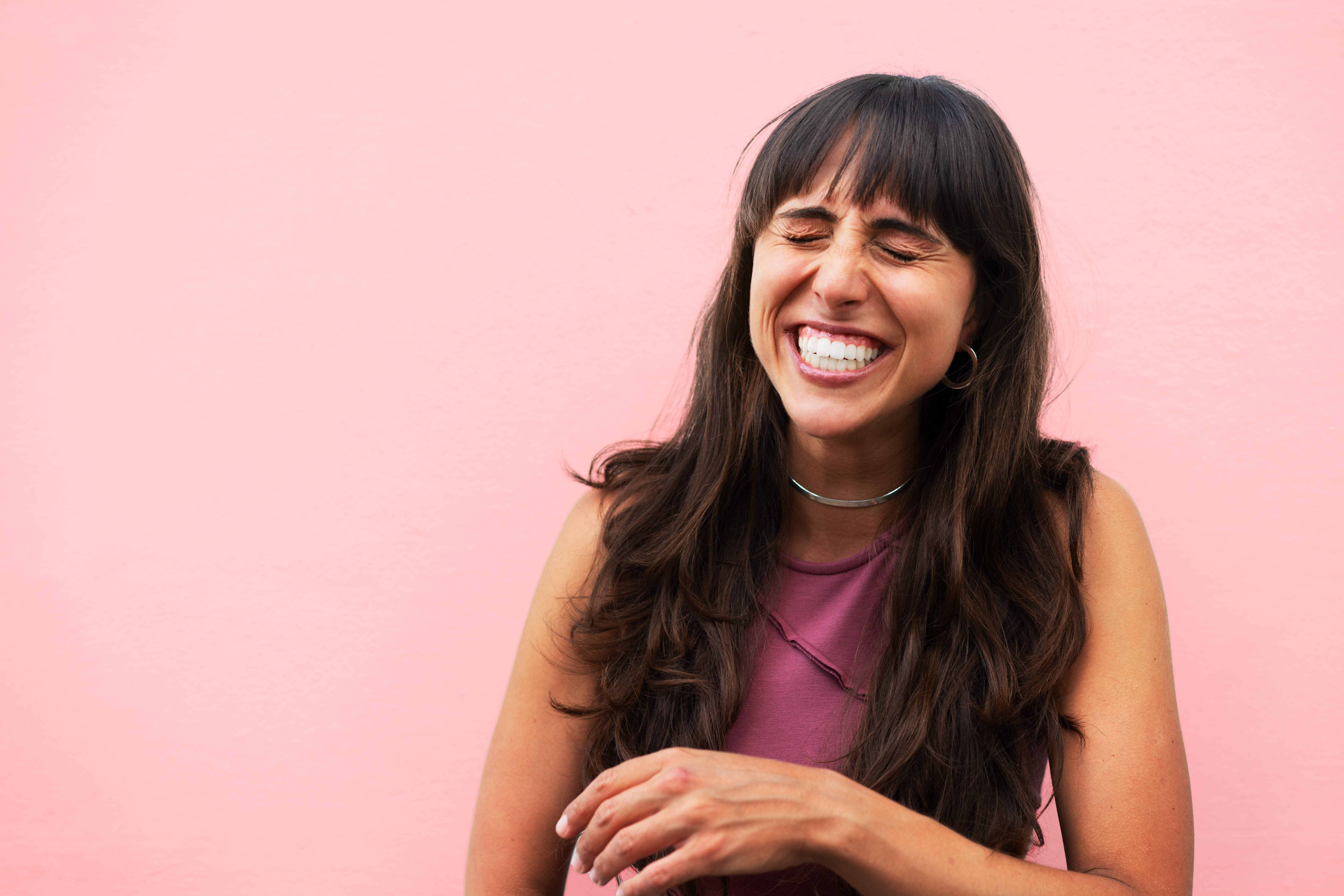 Person with agelong  hairsbreadth  laughing, eyes closed, against a pinkish  background, wearing a sleeveless apical  and necklace