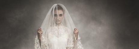 Person in vintage lace wedding dress and veil stands in foggy setting, holding the veil with both hands, gazing directly ahead