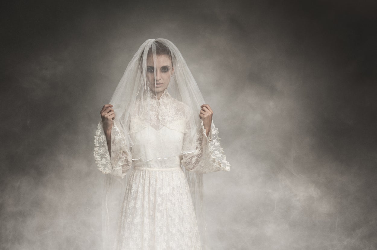 Person in vintage lace wedding dress and veil stands in foggy setting, holding the veil with both hands, gazing directly ahead