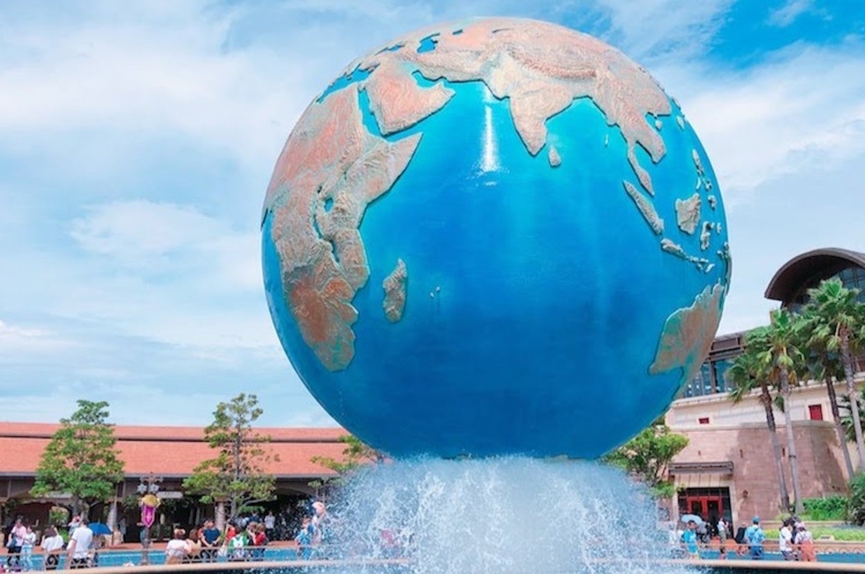 大きな地球儀の彫刻が噴水の上に設置され、多くの訪問者が周りを歩いている。背景に木々と建物が見える。