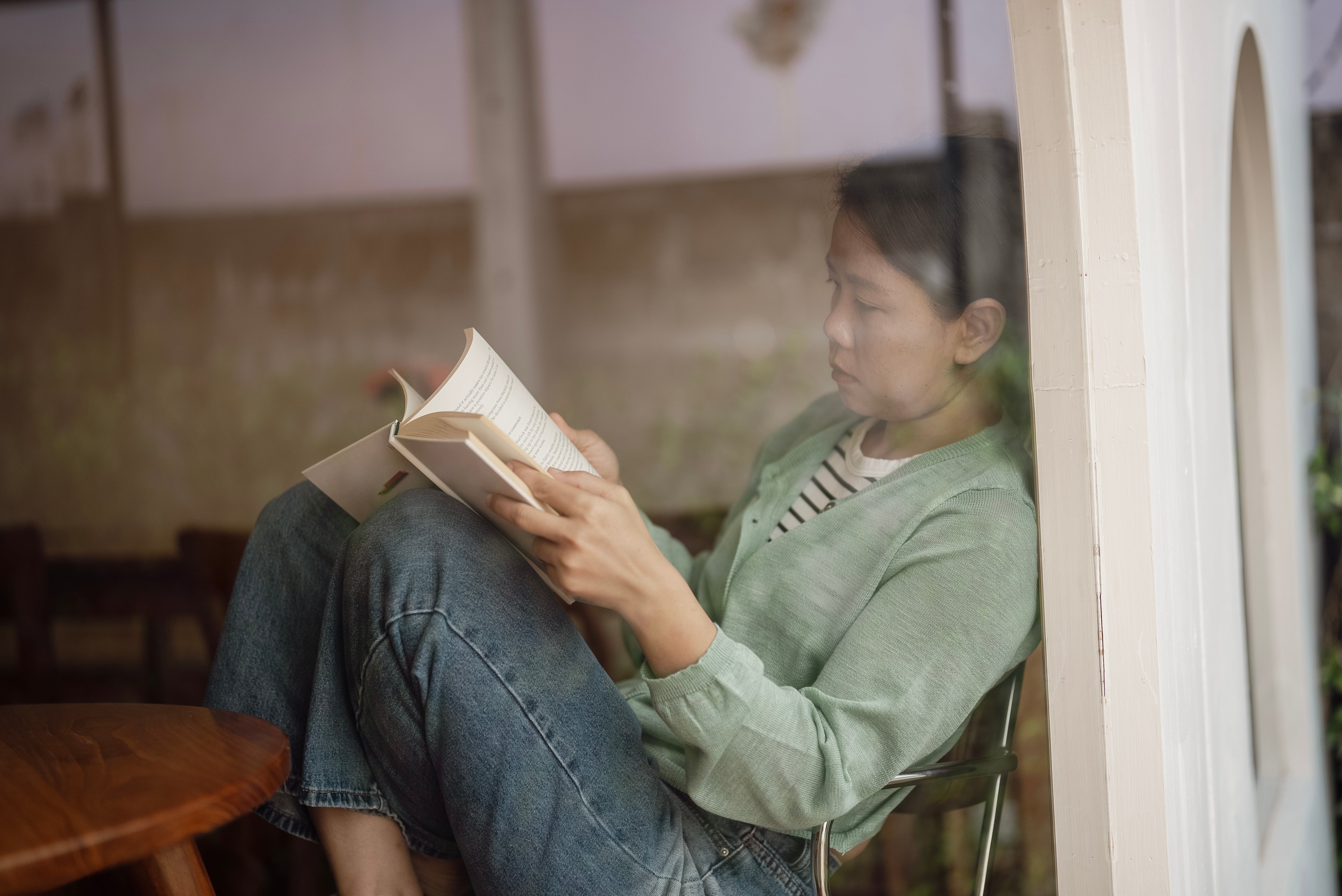 Person sitting by a window, reading a book. They wear a casual outfit consisting of jeans and a light sweater. Relaxed and focused