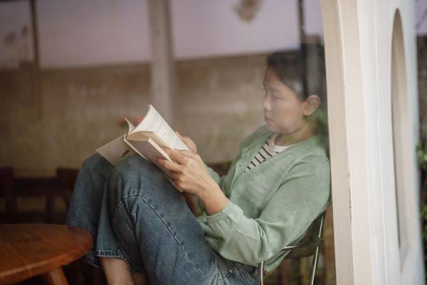 Person sitting by a window, reading a book. They wear a casual outfit consisting of jeans and a light sweater. Relaxed and focused