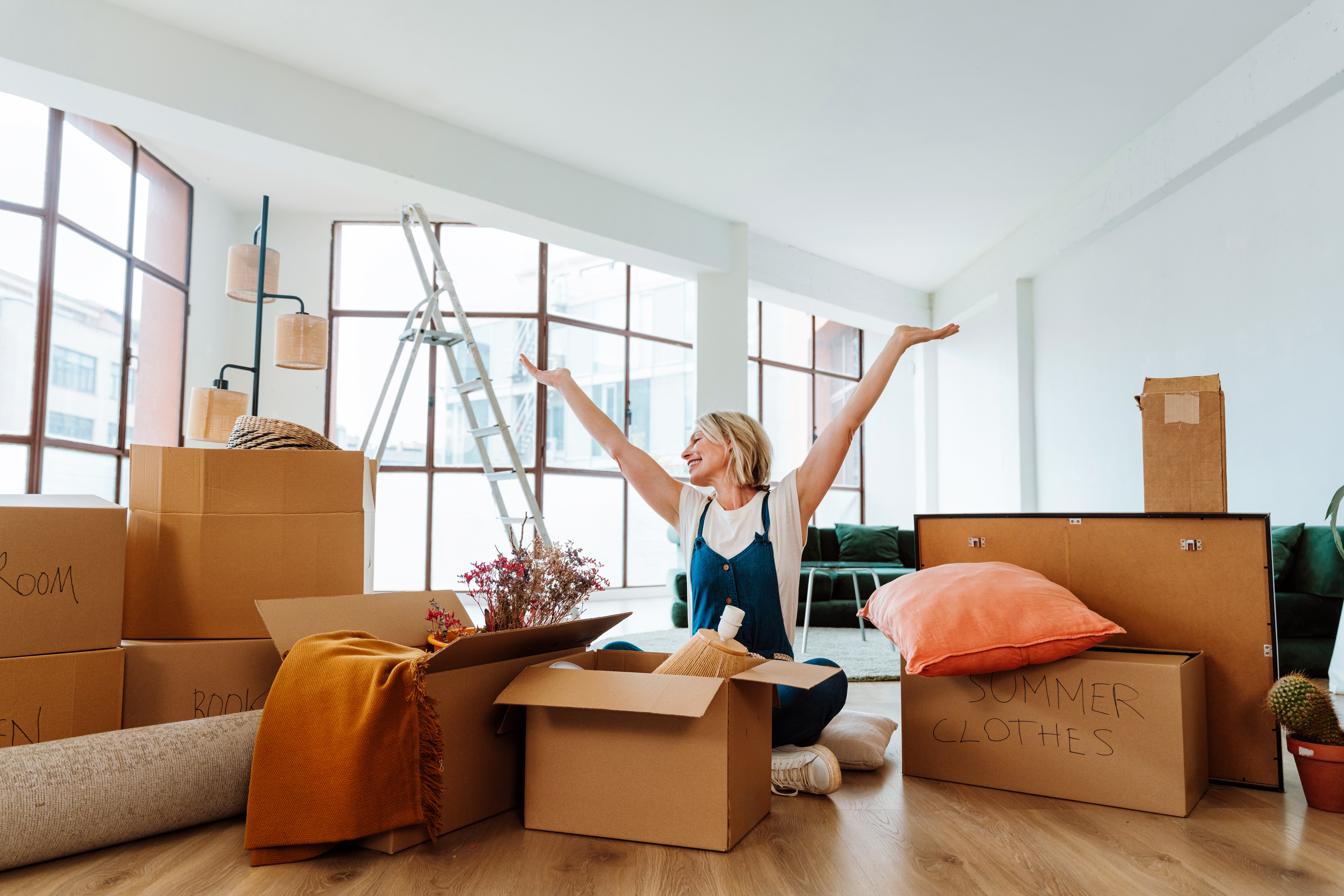 Woman happily unpacking moving boxes in a bright, spacious room with scattered home items and ladder in the background