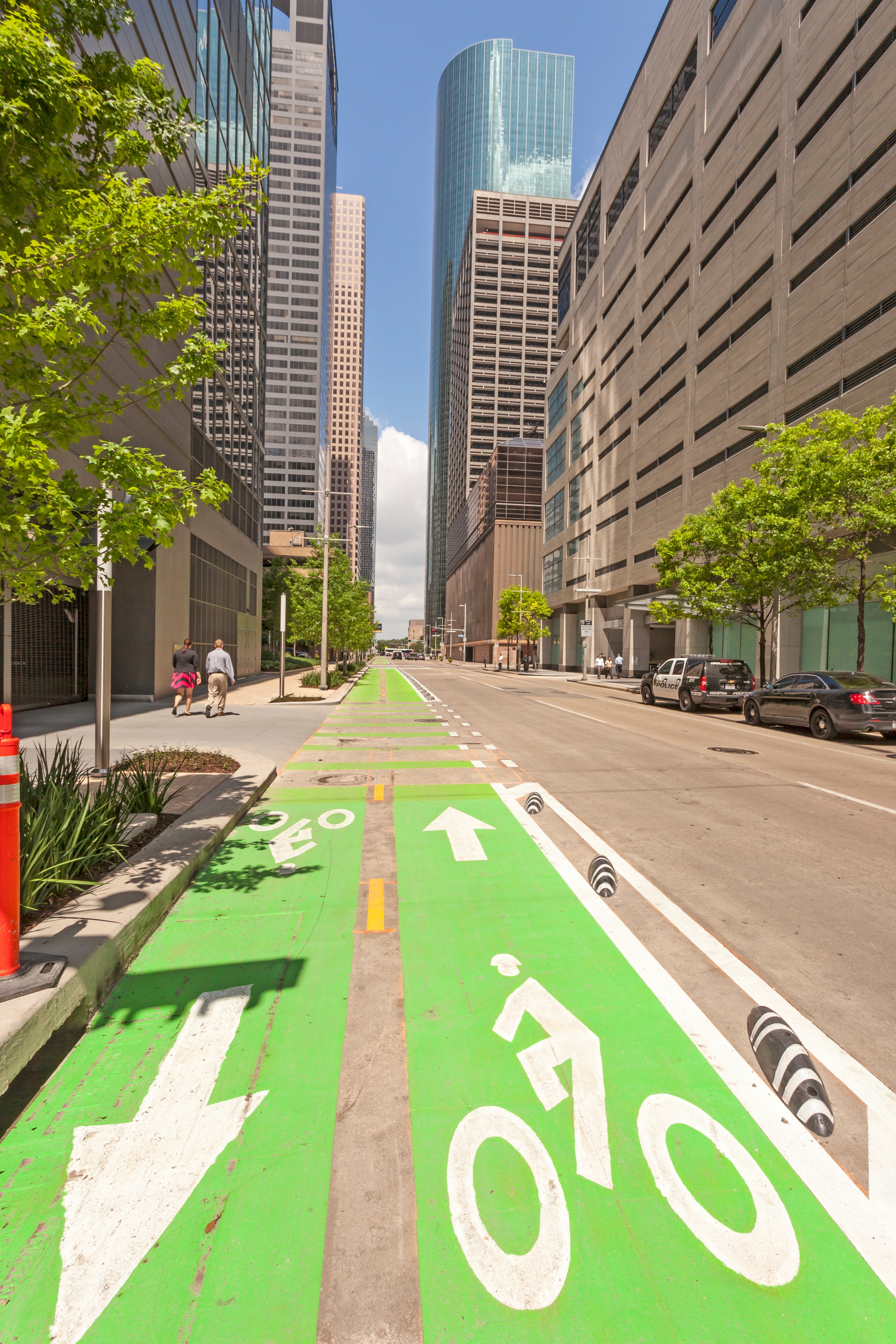 City street with a separated bike lane, arrow markings, and tall buildings on both sides. Pedestrians walk along the sidewalk