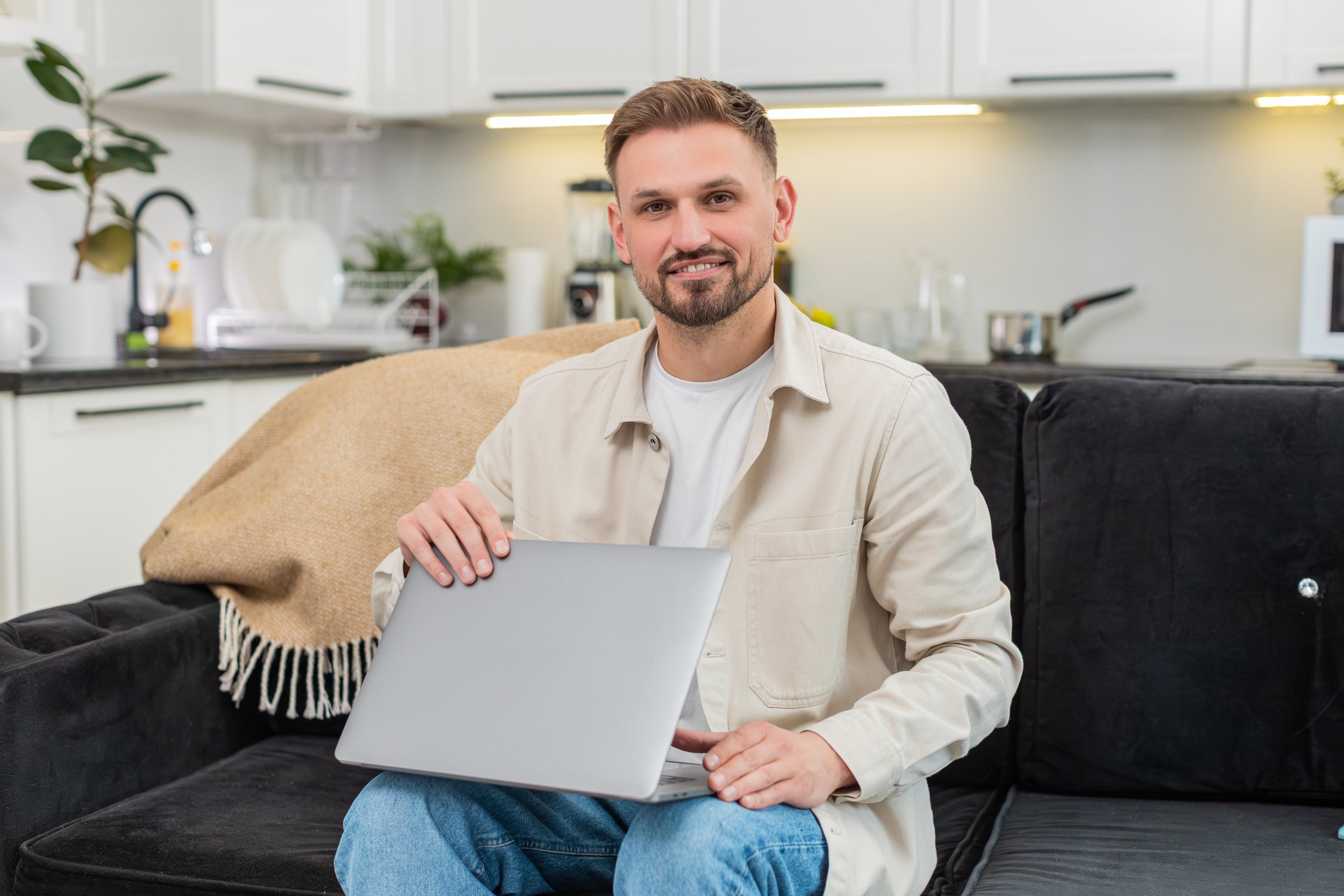 Person sitting on a couch in a modern kitchen holding a laptop and smiling. Casual attire includes a light shirt and jeans