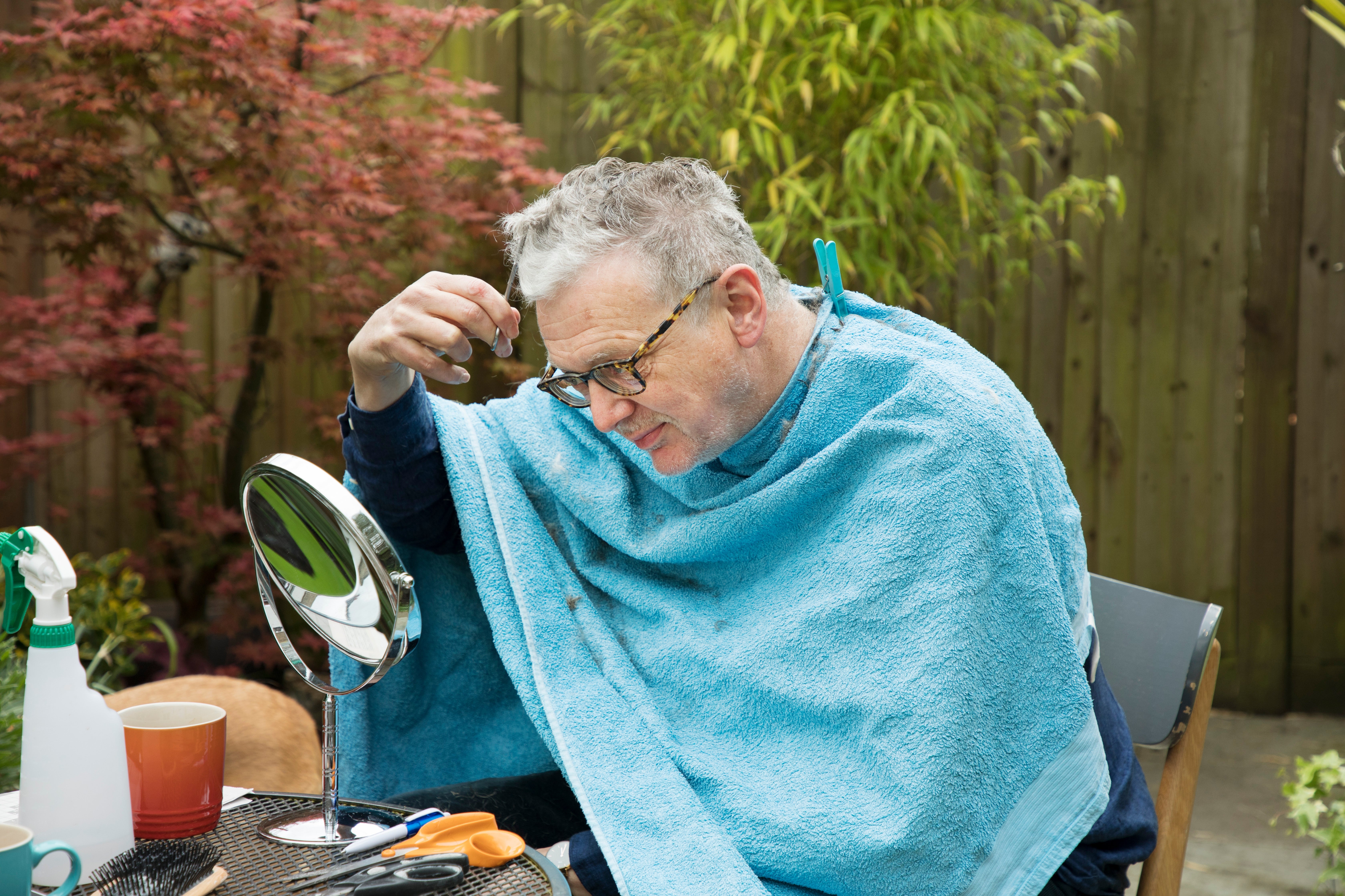 Man cutting his hair in a garden, using a mirror and scissors, with a towel draped around his shoulders