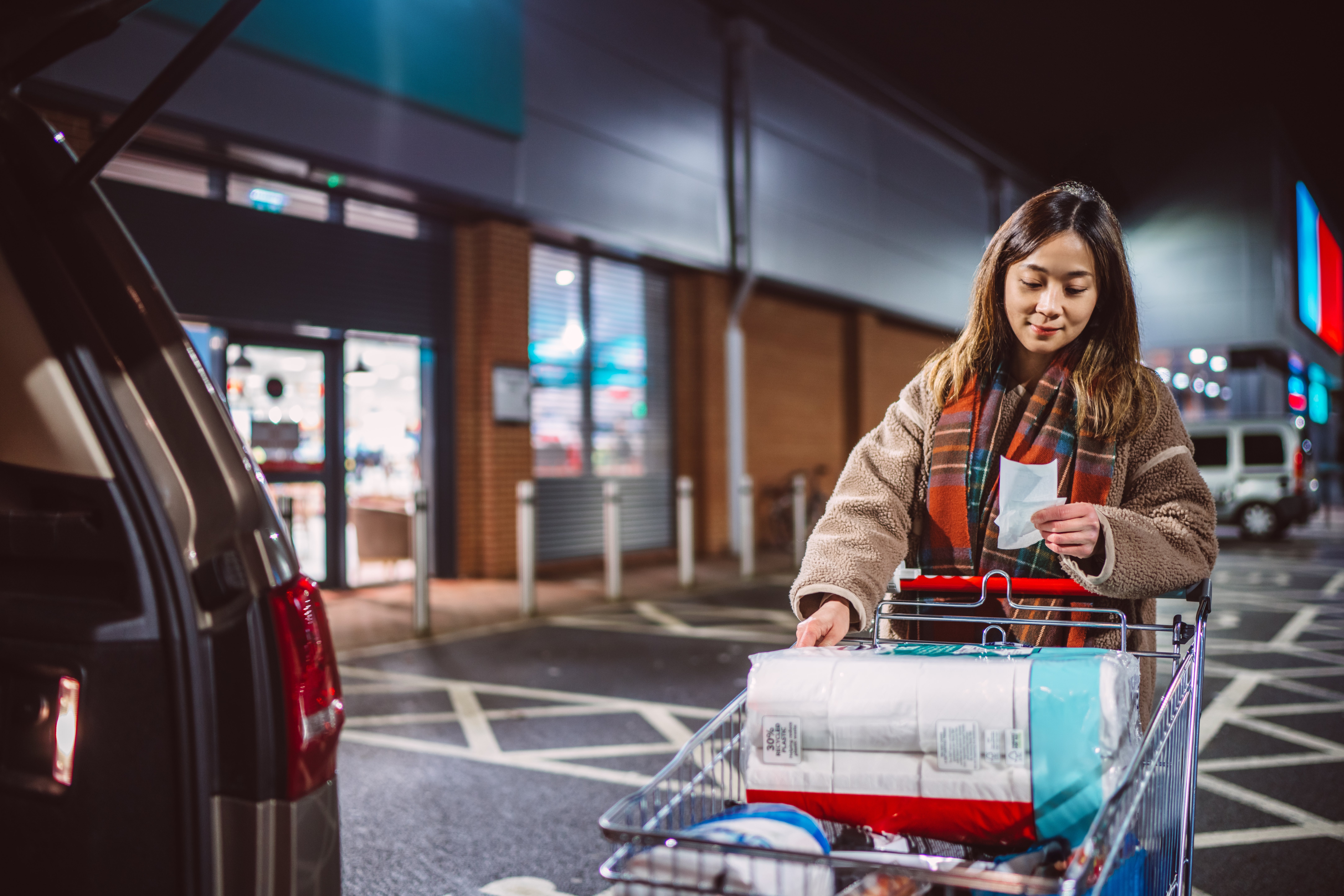 A person checks a shopping list while placing items into their car from a shopping cart in a parking lot at night