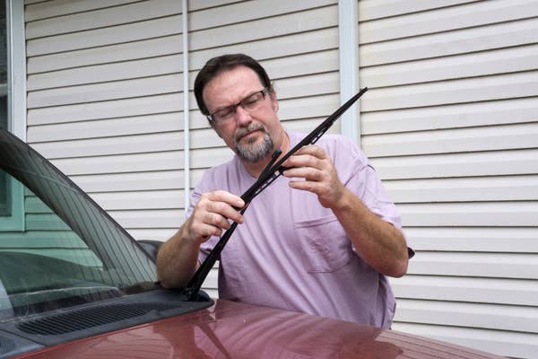 Man with glasses changes a car\&\#x27;s wiper blade, standing beside a vehicle parked in front of a house with siding