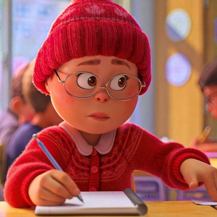 Un personaje animado con gafas y gorro rojo escribe en un cuaderno en un aula