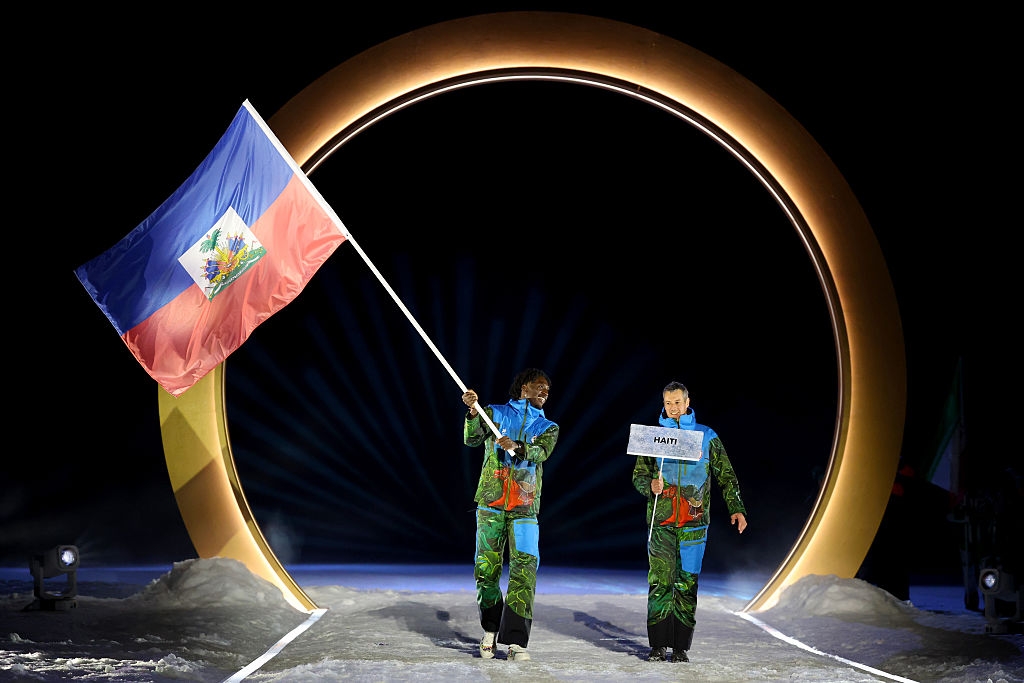 Two people in vibrant winter outfits walk under a large arch. One holds the Haitian flag, and the other holds a sign with "HAITI" written on it