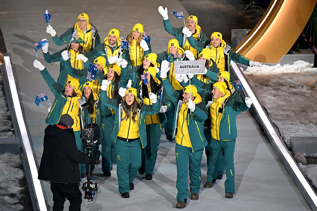 A group wearing winter sports uniforms waves flags at a sports ceremony. They smile, holding a sign that reads "Australia."
