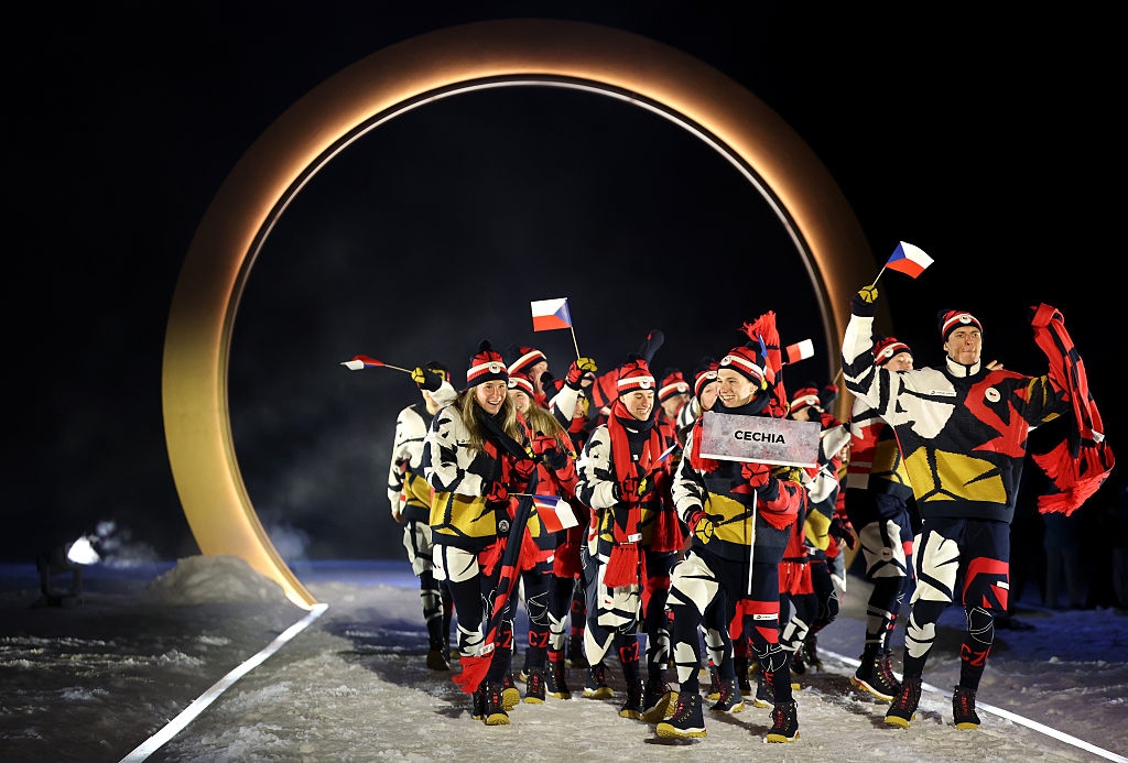 Athletes in patterned outfits hold flags and smile, walking under a lit arch at a winter event