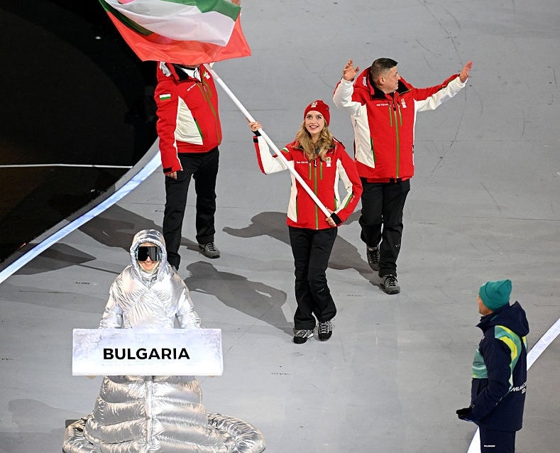 People march in a sports event. A person holds the Bulgarian flag, flanked by others in red and black winter attire. A sign reads "Bulgaria."