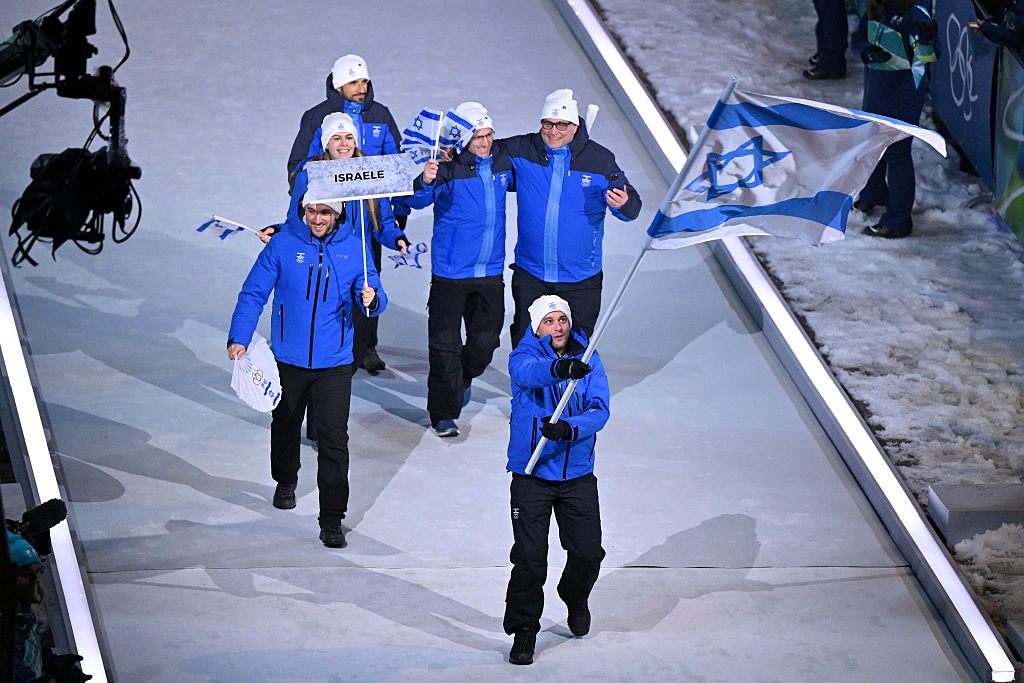 Athletes in blue jackets, wearing hats, march with Israeli flags during a winter sports ceremony on an ice rink
