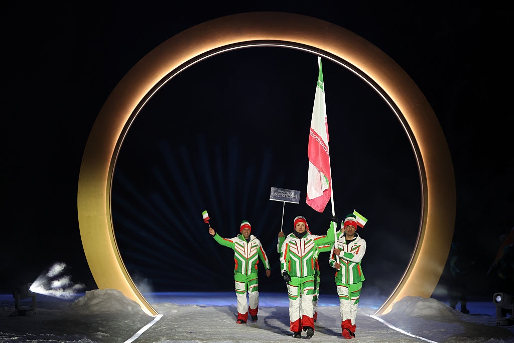 Three athletes in matching outfits parade with a flag and sign at a ceremonial event, under a large illuminated arch