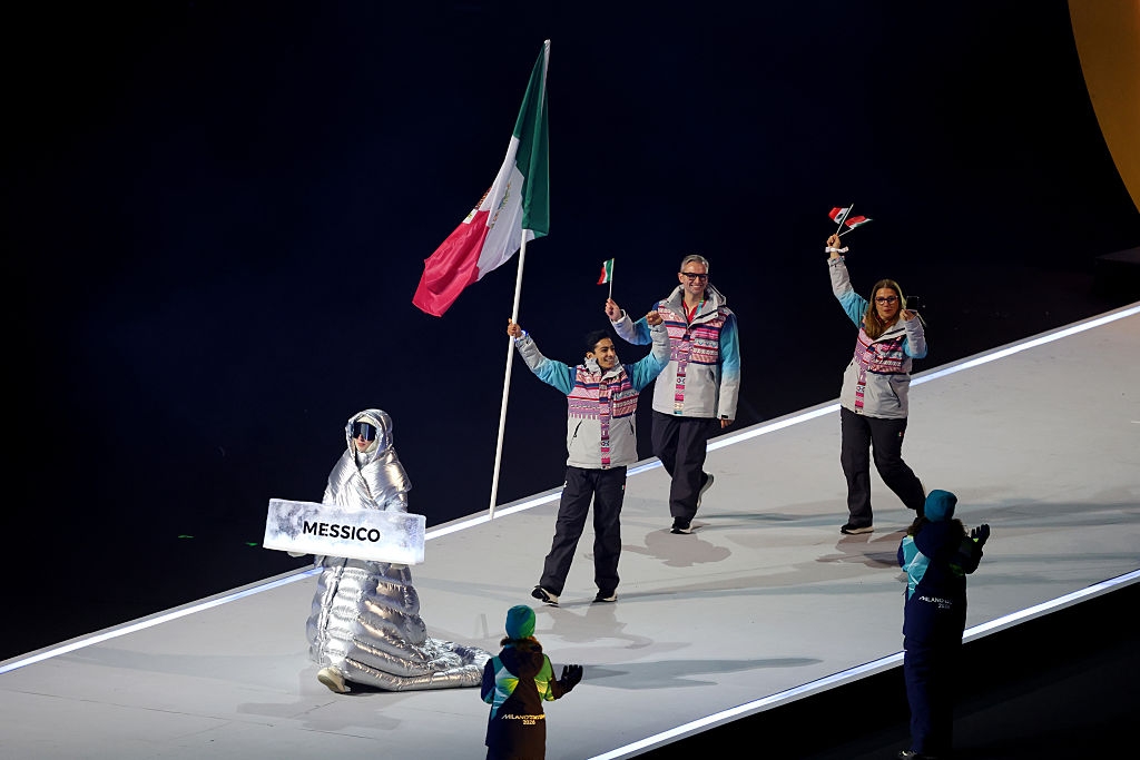 Person in silver cloak leads Mexico's team with flag at ceremony, followed by three individuals in patterned jackets on stage