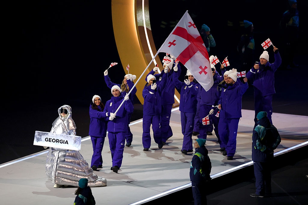 Parade team carrying Georgia flag, wearing blue winter gear and hats, led by a person in a metallic suit holding a "Georgia" sign