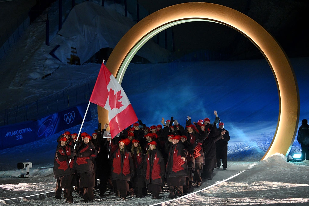 Athletes in winter sports uniforms wave as they carry a Canadian flag, entering through a large circular structure at an outdoor event