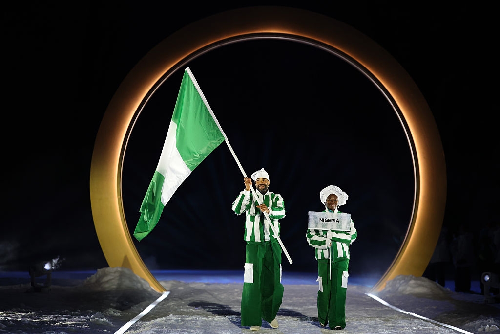 Two people in matching outfits hold a large flag with a vertical green, white, green design on a snowy path under a glowing circular arch