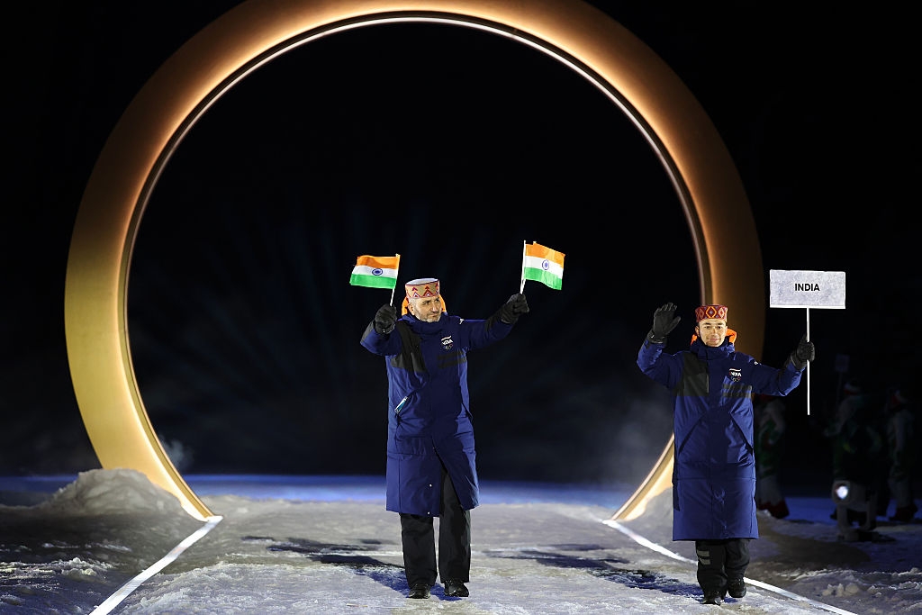 Two people in winter coats wave flags under an illuminated arch at a nighttime event, one holding a sign that reads "India."