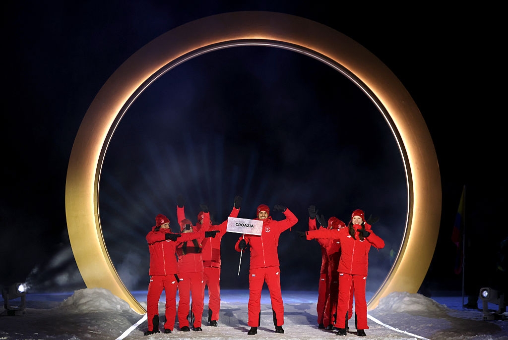 Group of athletes in matching red outfits, holding a "Croatia" sign, walk through a large illuminated ring at a nighttime event