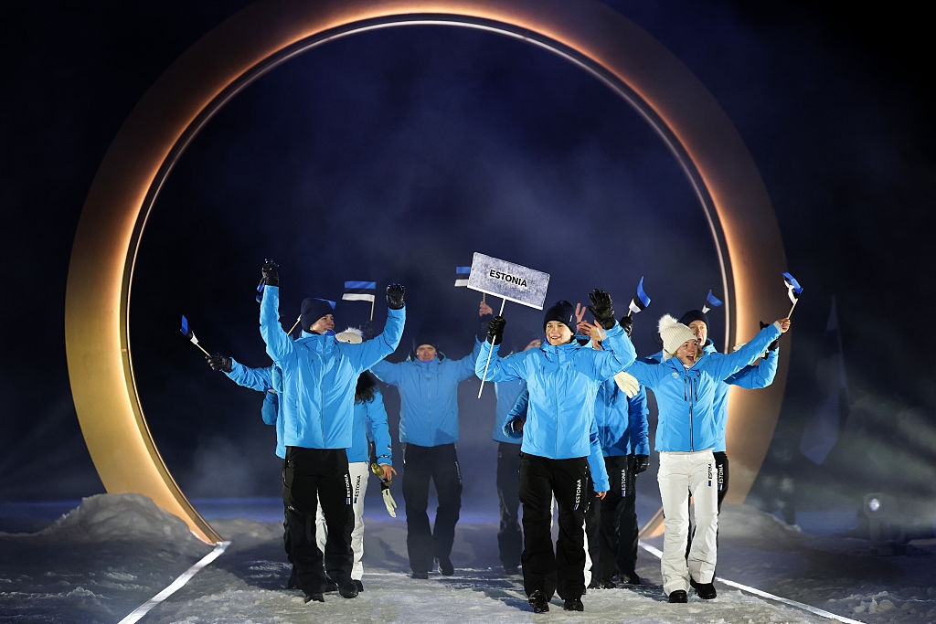 A group of people in matching winter jackets wave flags, walking enthusiastically through a circular archway at an event