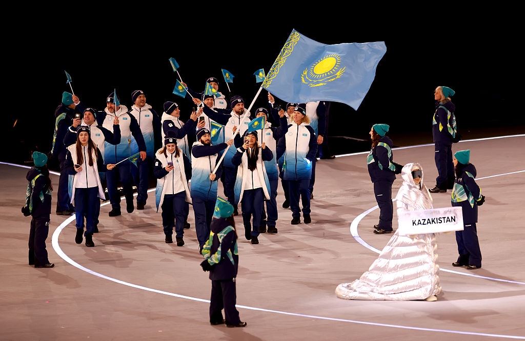 Kazakhstan's team enters a stadium during an event, with members wearing matching warm outfits and waving flags. A person at front holds a "Kazakhstan" sign