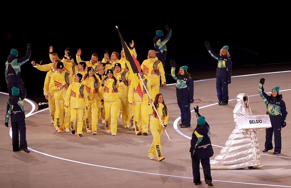 Belgian athletes in yellow tracksuits wave at an event parade, led by a sign holder in a silver puffy outfit with "Belgium" on the sign