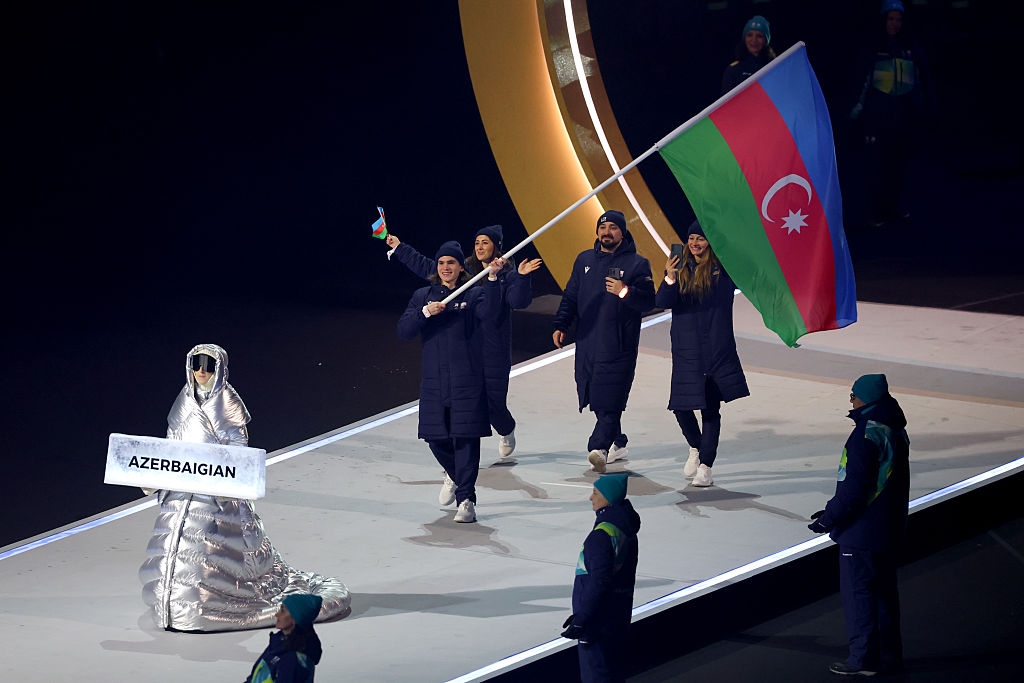 Person in shiny silver outfit leads Azerbaijan's Olympic team, who are in dark coats, holding a flag during a parade