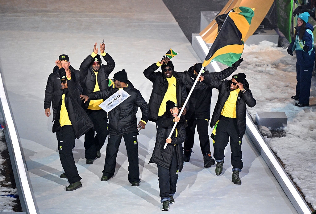 Team in matching winter gear, including hats and jackets, proudly waves a Jamaican flag at an outdoor event, walking on snowy ground