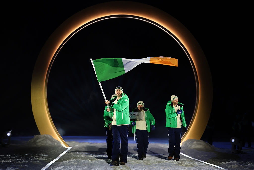 Group carrying Irish flag walks through illuminated ring during an outdoor event. Participants wear green jackets and hats