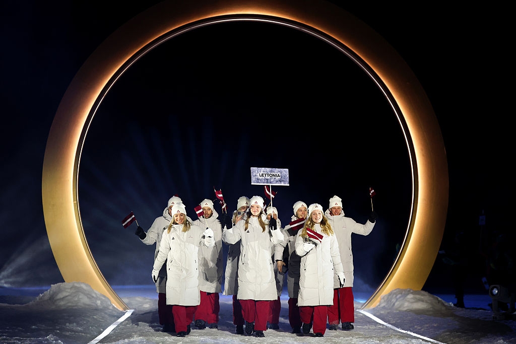 Group of people in matching winter outfits carrying flags, walking through a large circular structure, under a "Latvia" sign, on a snowy pathway