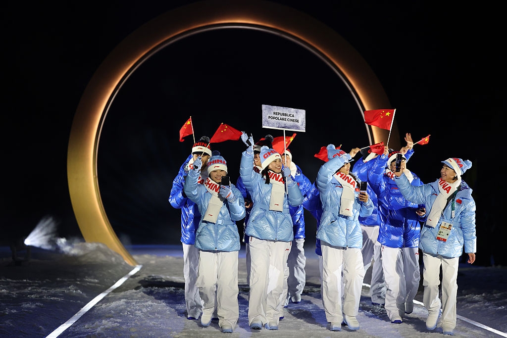 Athletes in winter gear hold flags and a sign for the Paralympic Games during a ceremony under a large circular arch structure
