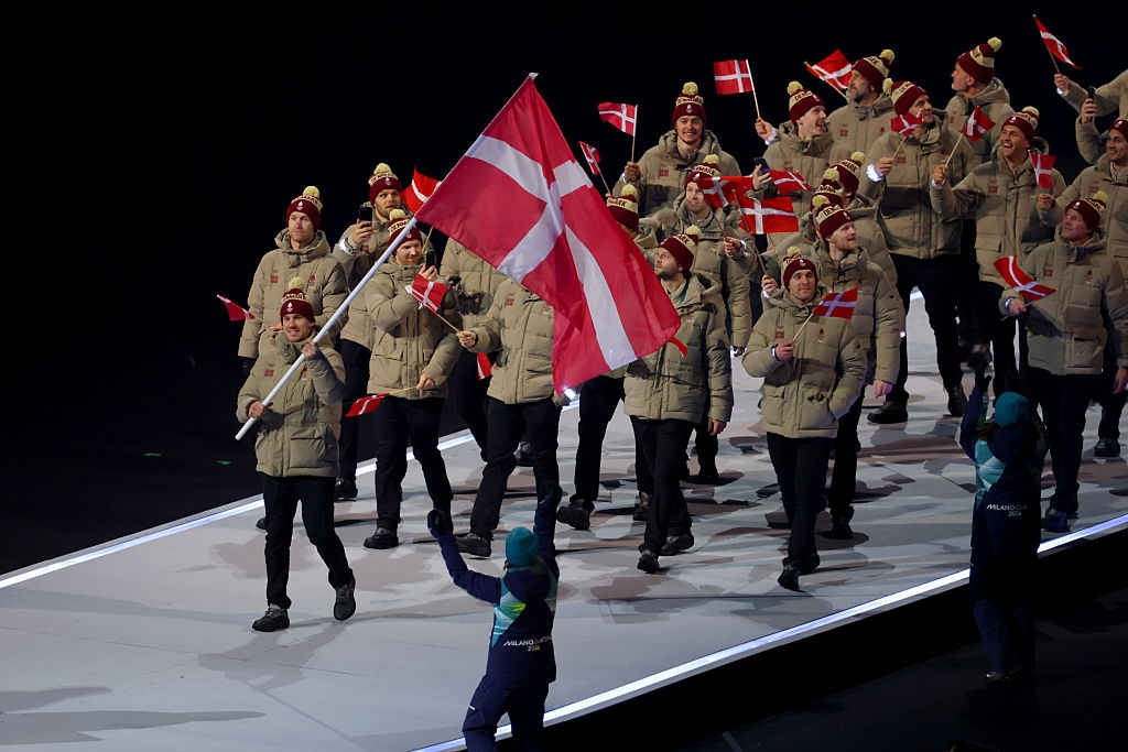 A team of athletes in tan winter uniforms march with a large Danish flag during an Olympic opening ceremony, waving smaller flags