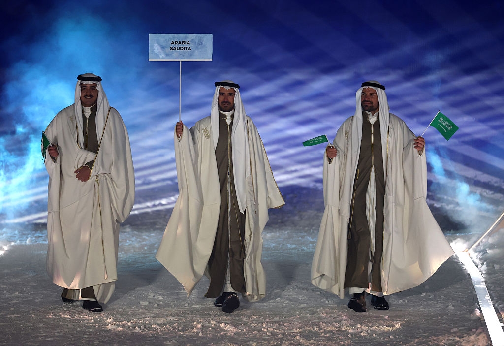 Three men in traditional attire hold signs and flags, walking on a snowy path during a ceremony