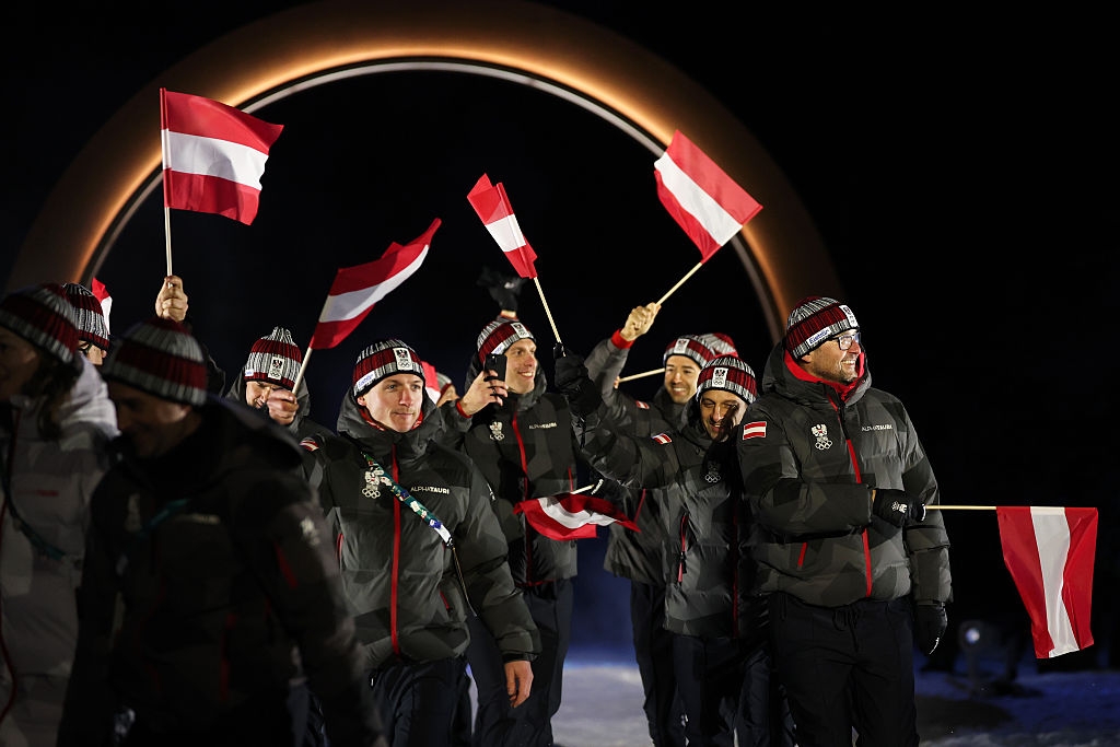 Group of athletes in winter gear joyfully wave flags during a ceremony, standing under a large, illuminated arch