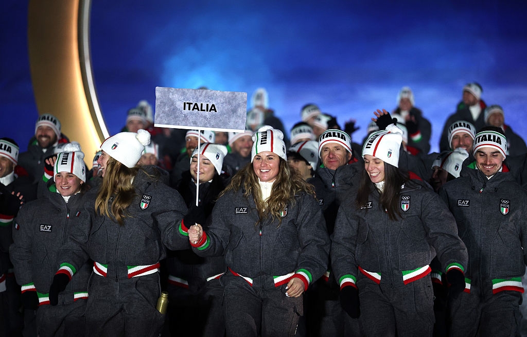 Group of Italian athletes in uniform and beanies, smiling and waving, holding an "Italia" sign at an Olympic ceremony