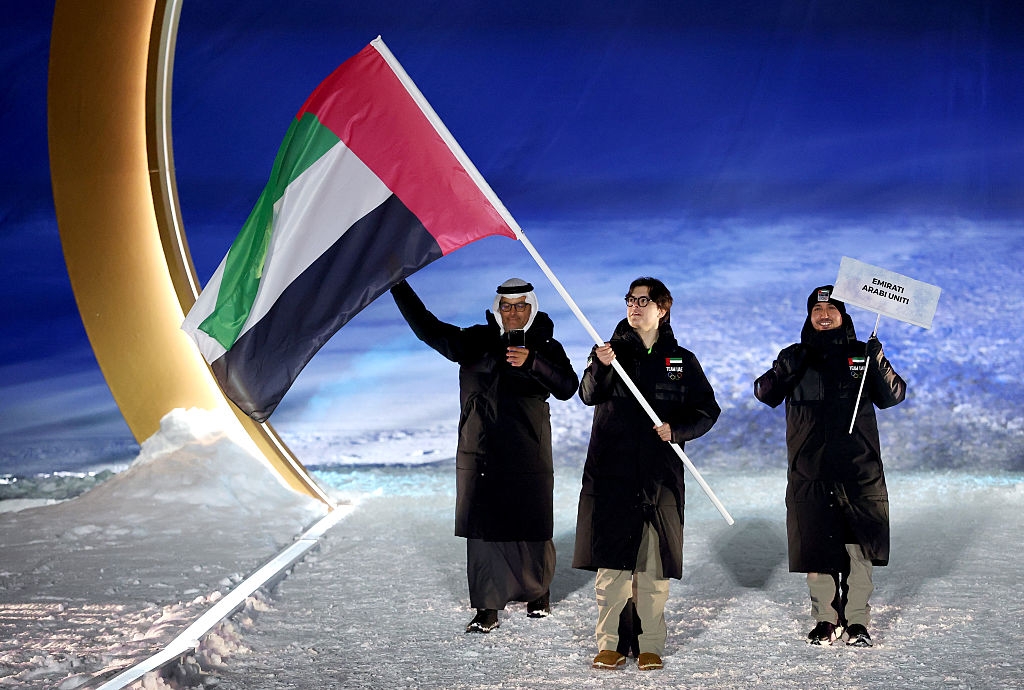 Individuals in winter attire hold UAE flag and a sign reading "Winter Games". They walk on a snowy surface at a wintry-themed event