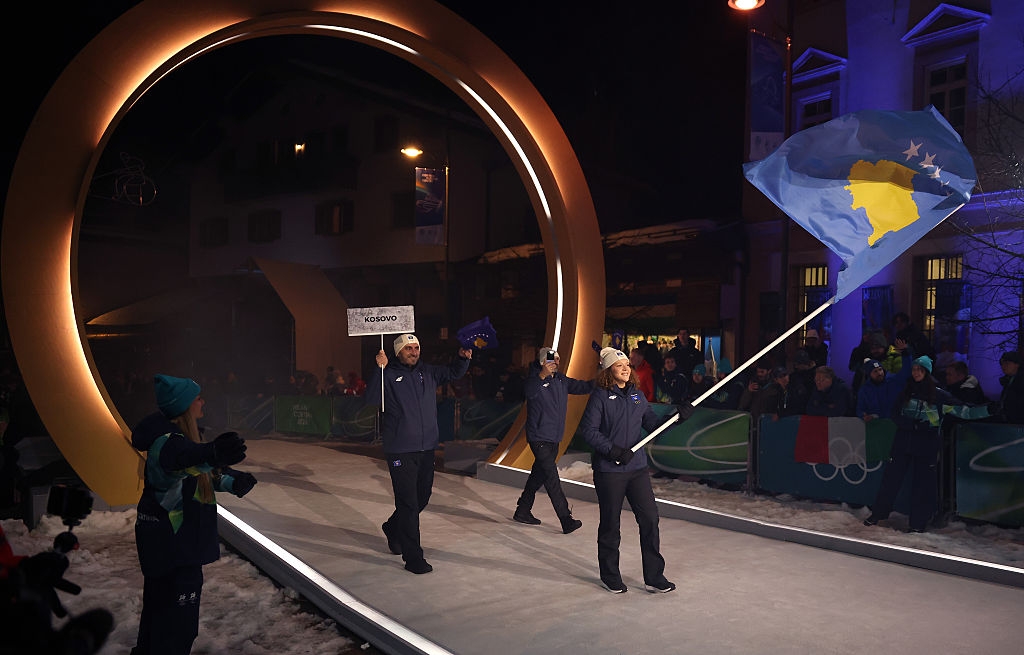 People in winter sportswear hold a national flag and a sign. They walk under a large illuminated ring outdoors at a nighttime event