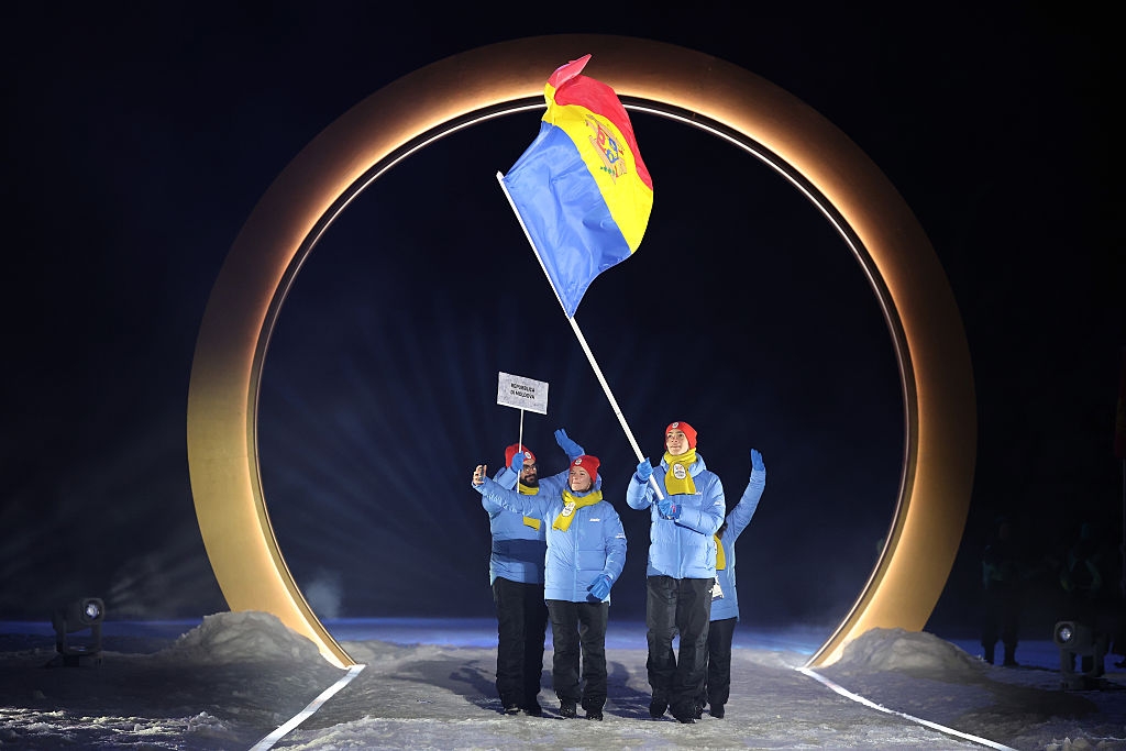 Four people in blue jackets wave a flag and hold a sign, walking through a lit circular arch at a ceremony