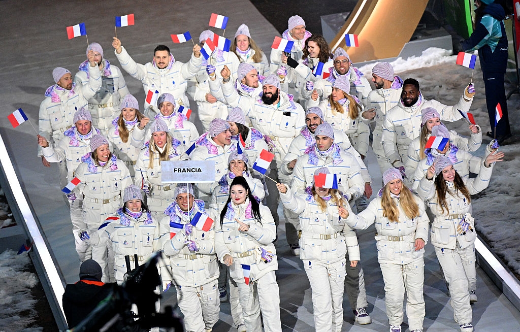 Group of athletes in winter gear carrying French flags in a parade, marked with a "Francia" sign, on a sloped platform, snowy background