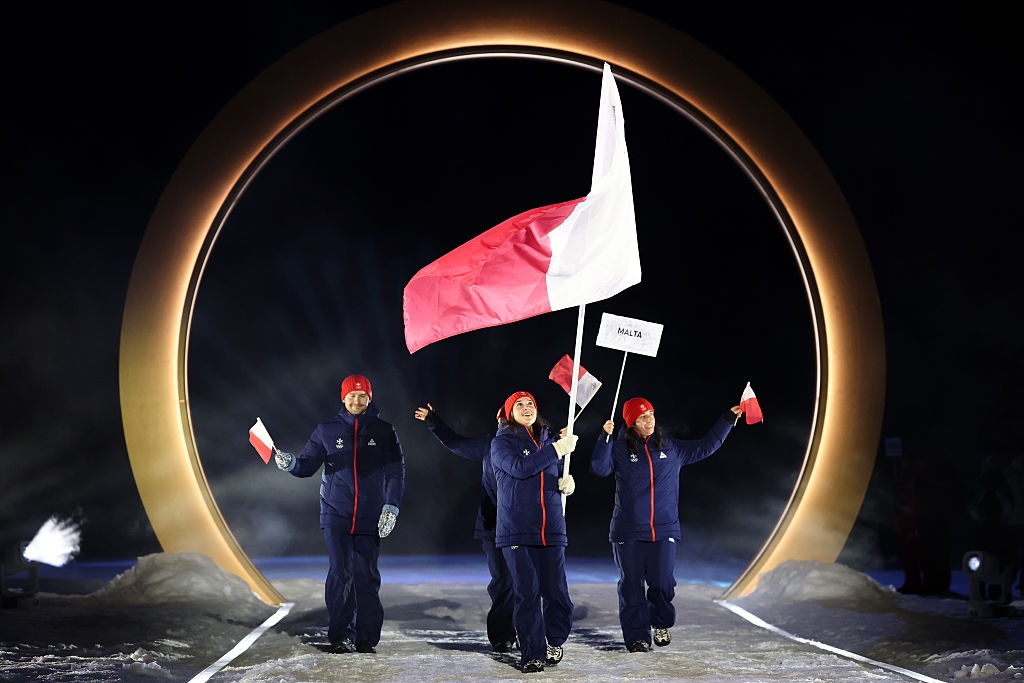 People wearing winter gear walk through a large circular arch, carrying a flag and small signs