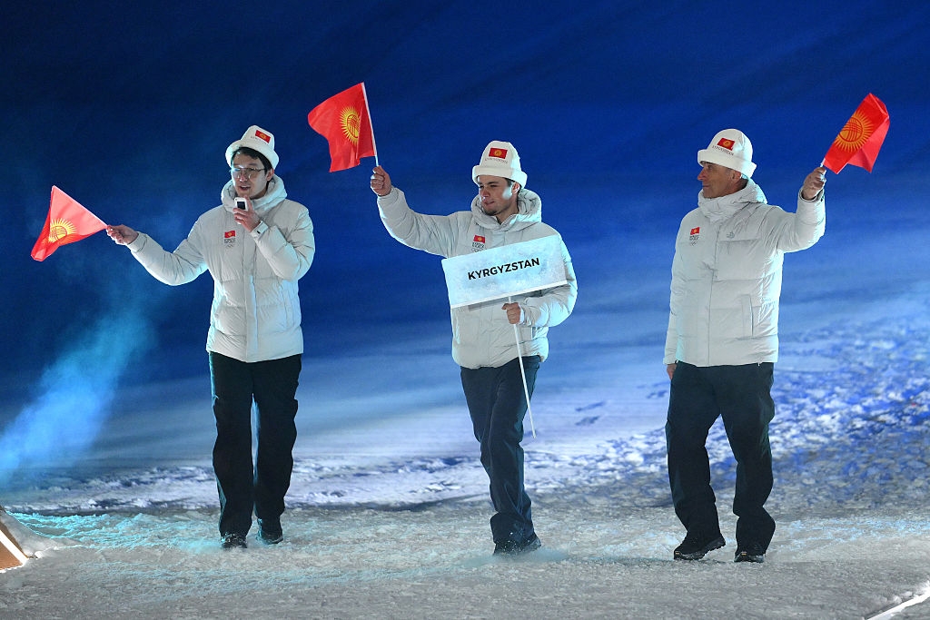 Three people in winter clothing wave Kyrgyzstan flags and hold a sign labeled "Kyrgyzstan," walking on a snowy surface
